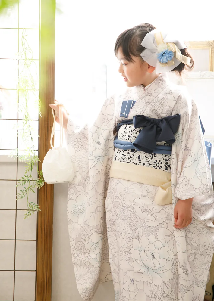 白い花柄の着物に紺の帯を締め、巾着を持って障子の窓辺に立つ七五三のお子さま