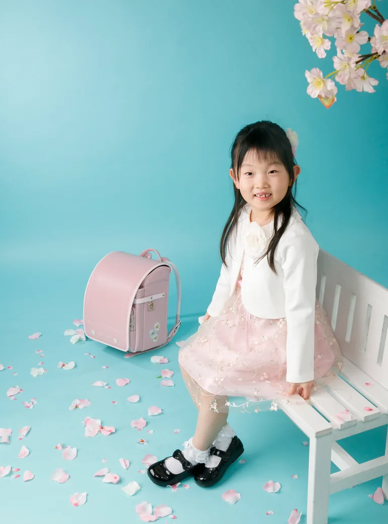 桜と水色の背景の前で、ランドセルを置いた白いベンチに腰かける入学前のお子さま